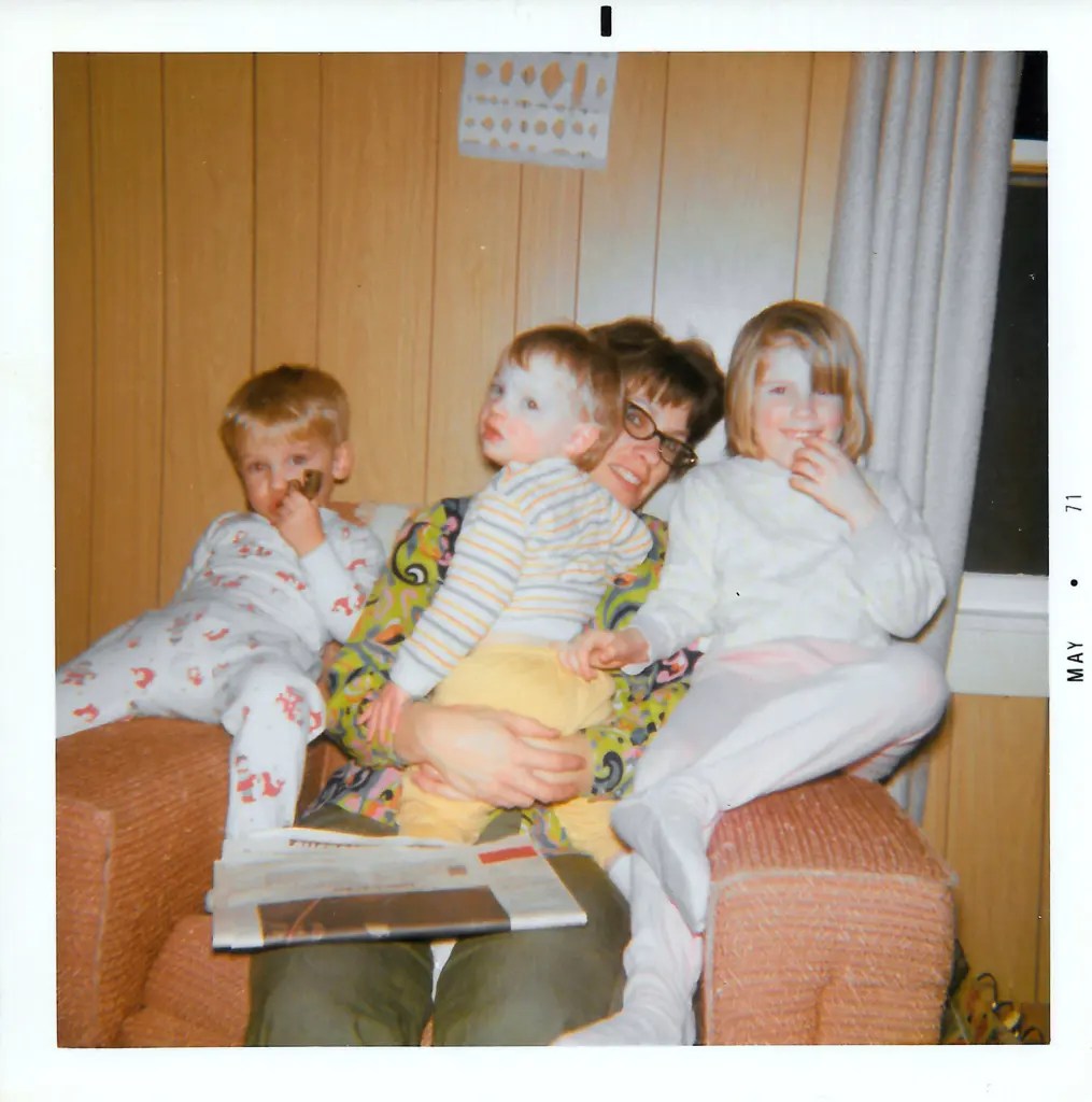 A person wearing glasses and three children sit close together  in an armchair with a newspaper on the person's lap in a wood-paneled room.