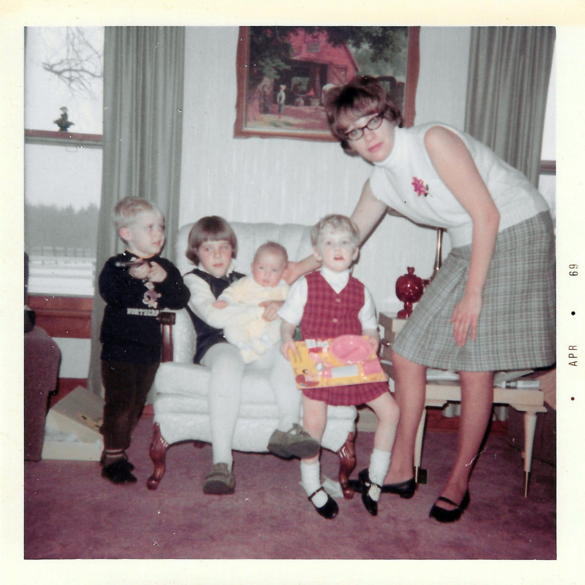 An adult person stands beside four children in a room, with one child holding a baby in a chair and another holding a toy. Behind them are framed art and curtains on windows.