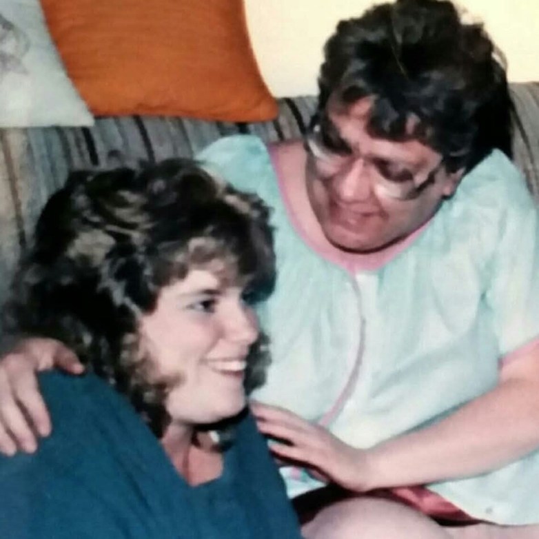 Two people sit close together and smile on a couch, with one person’s arm around the other.