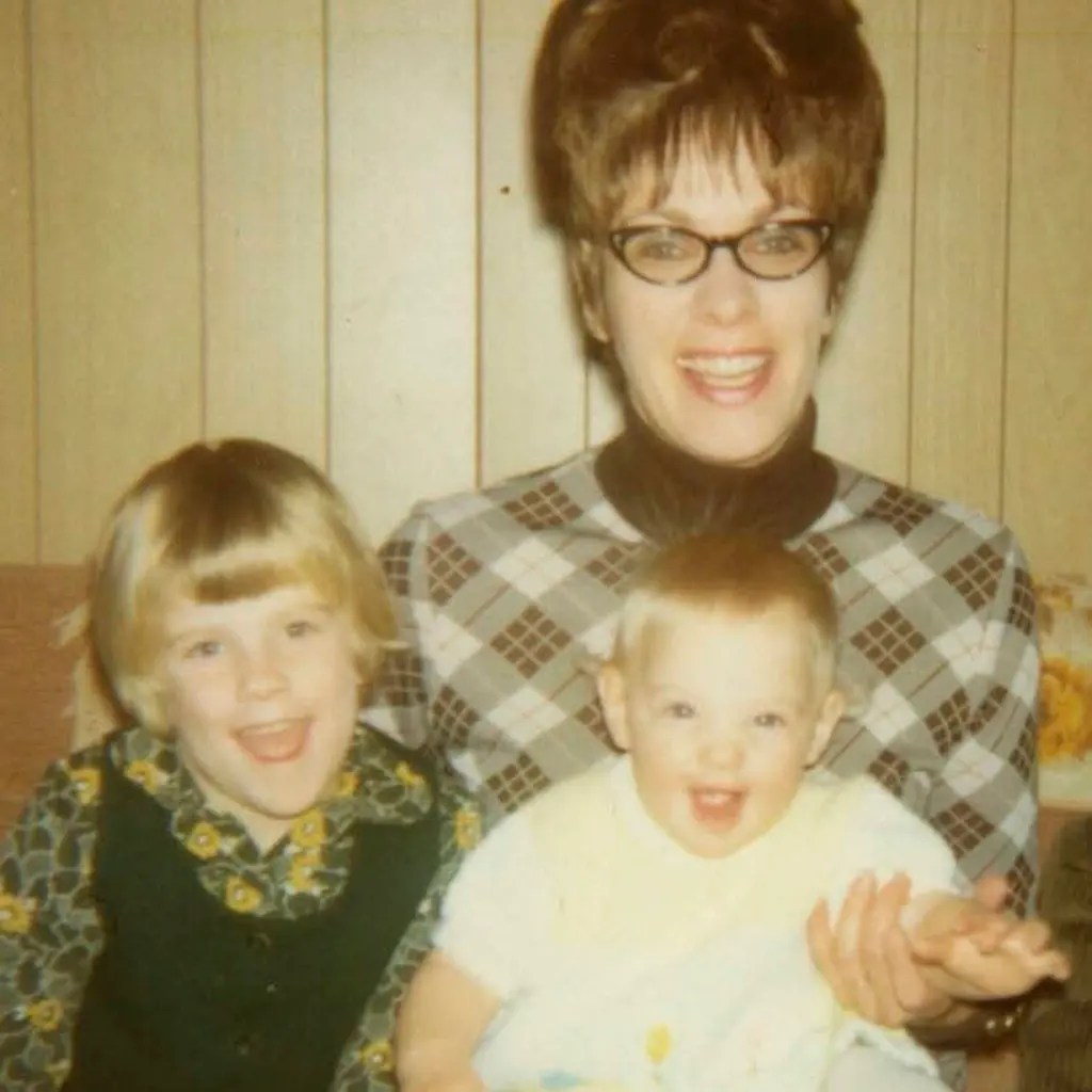 A person wearing glasses sits with two children, all smiling in front of a wood-paneled wall.