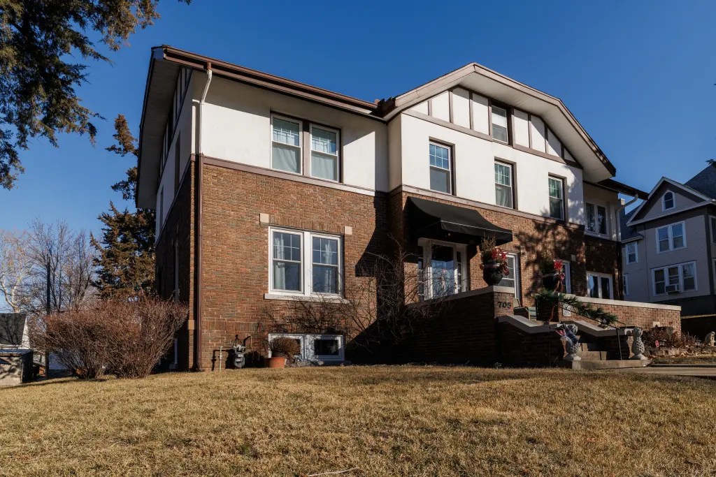 A two-story brick house with white trim and a black awning over the front door, with a lawn in front and steps leading up to the entrance. Other homes are nearby.