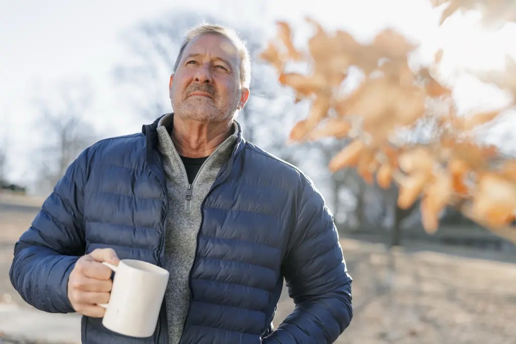 A person wearing a blue jacket holds a white mug outdoors, with bare trees and autumn leaves visible in soft focus.