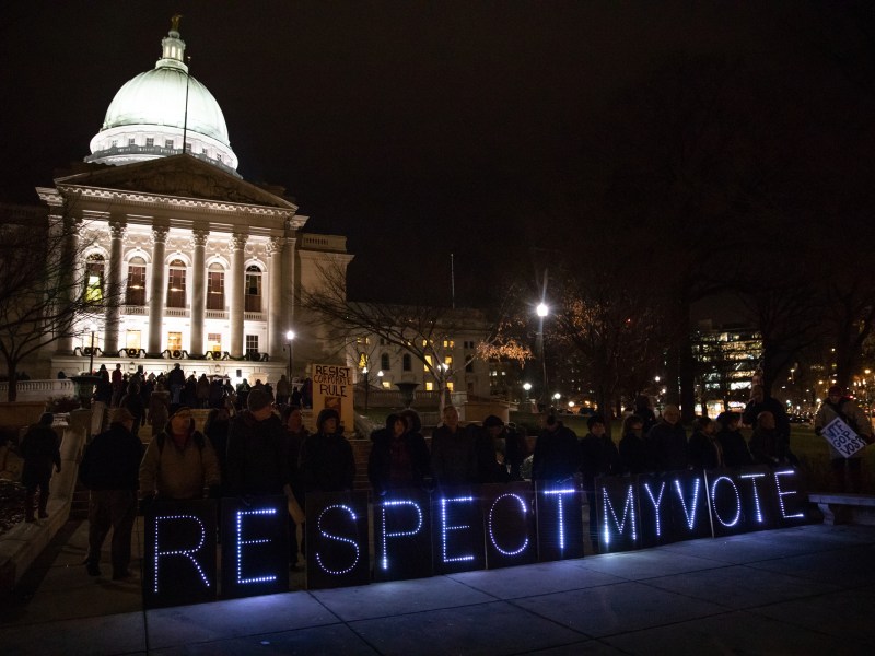 People gather at night outside a lit domed building with illuminated letters spelling “RESPECT MY VOTE” next to a sidewalk.