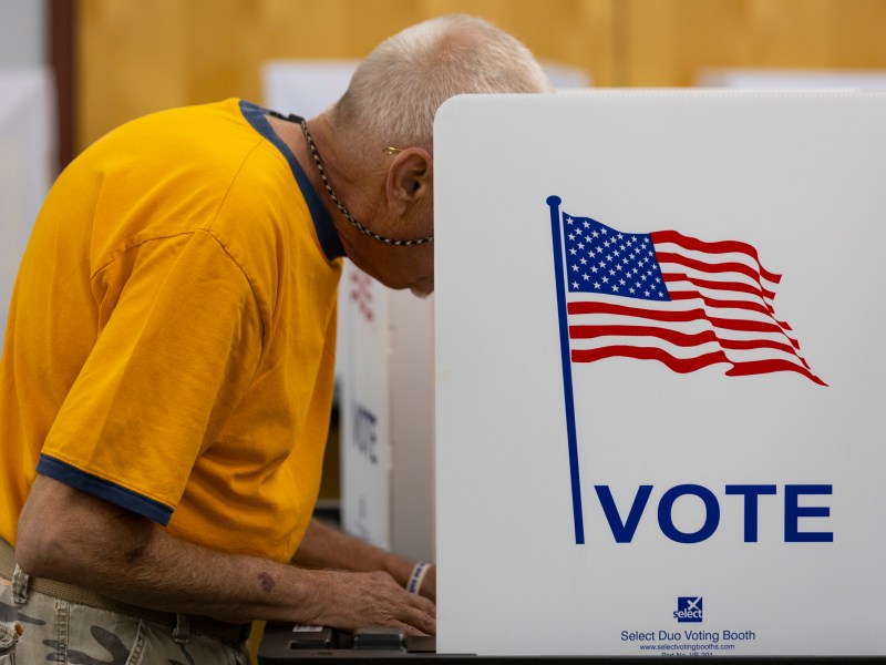A man in a yellow shirt stands next to a "VOTE" sign with an American flag.