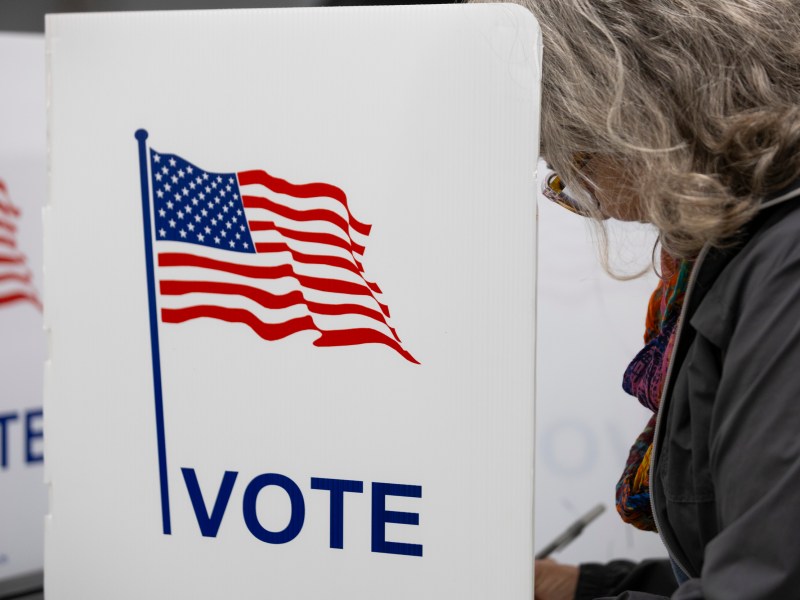A person holds a pen and stands at a white voting booth marked with a U.S. flag graphic and the word “VOTE”