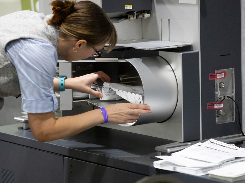 A person reaches into a machine with perforated paper sheets inside it while additional sheets are stacked on the counter nearby.