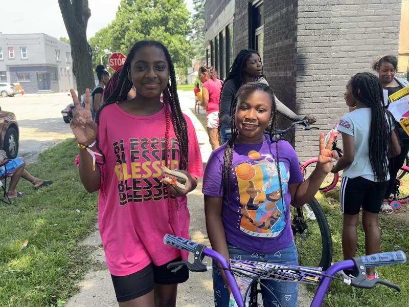 Two girls smile and each hold up two fingers and stand near a bike.