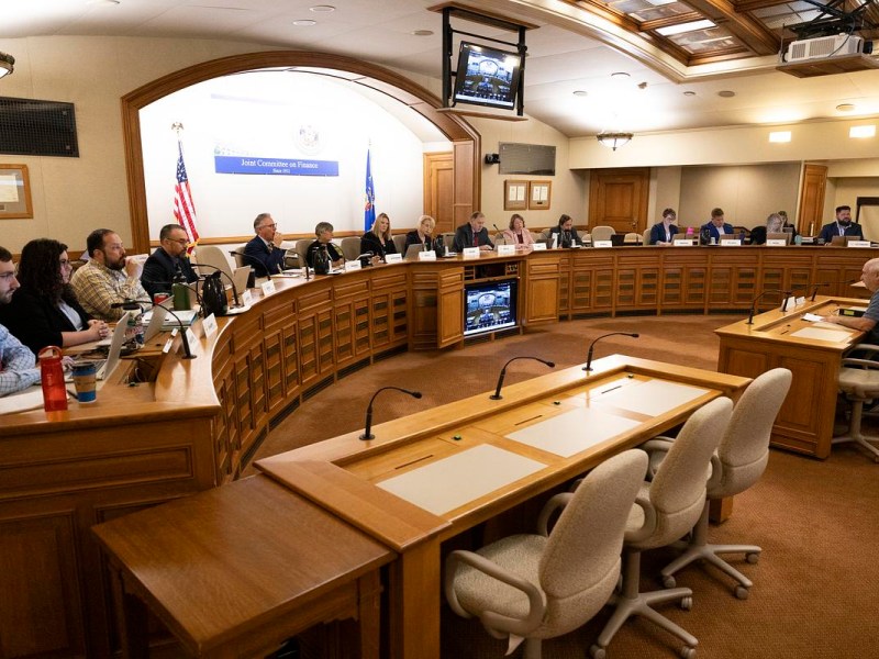 People sit around curved desks in a hearing room with microphones, laptops, and monitors, facing a central table beneath a sign reading "Joint Committee on Finance"