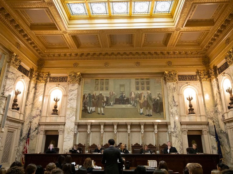An ornate room with marble columns and a high ceiling with a skylight features several people seated behind a large bench while a person stands and others are seated facing them.