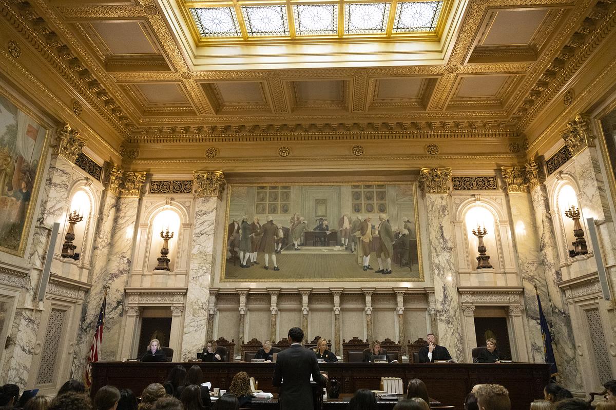 An ornate room with marble columns and a high ceiling with a skylight features several people seated behind a large bench while a person stands and others are seated facing them.