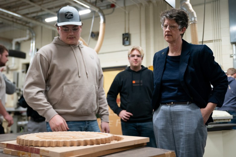 Three people wearing safety glasses stand around a wooden gear-shaped piece on a table in a large room with machinery and ventilation ducts visible and other people in the background.