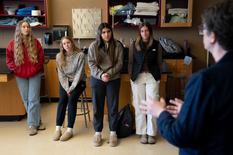 Four people stand and sit in a room with cabinets, drawers, a sink and other items behind them, looking at a person who is gesturing in the foreground.