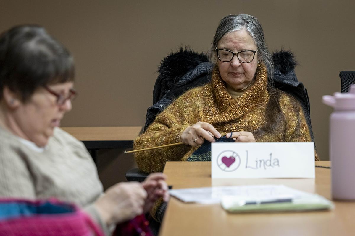 A person knits with needles at a table, with a name card reading “Linda” and papers and a water bottle nearby, while another person also knits at the table.