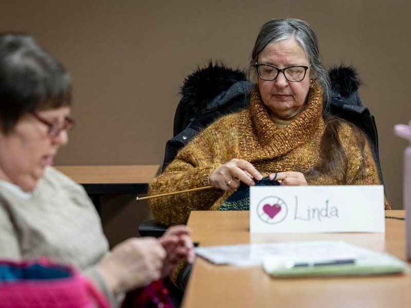 A person knits with needles at a table, with a name card reading “Linda” and papers and a water bottle nearby, while another person also knits at the table.