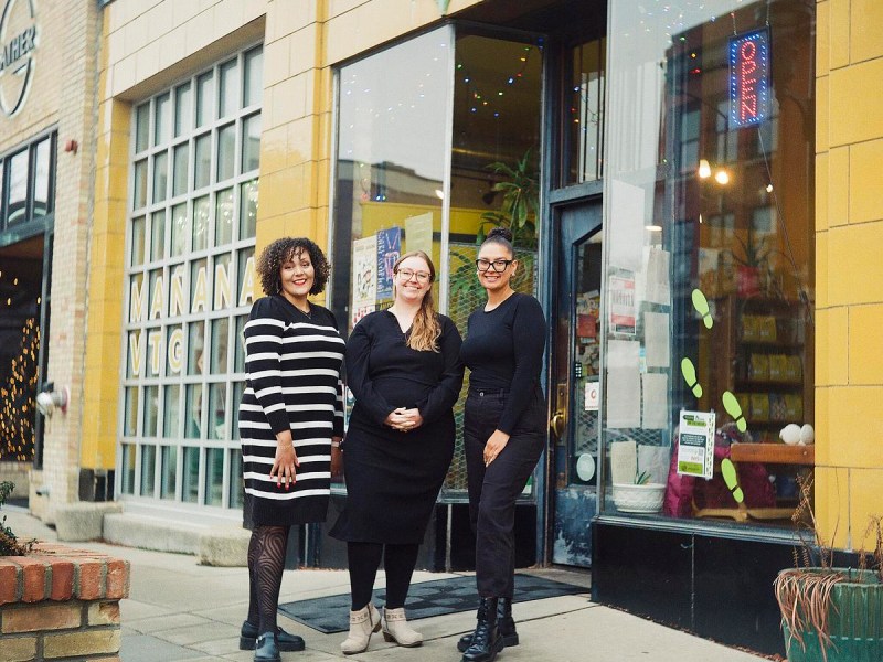 Three women stand together and smile outside a storefront.