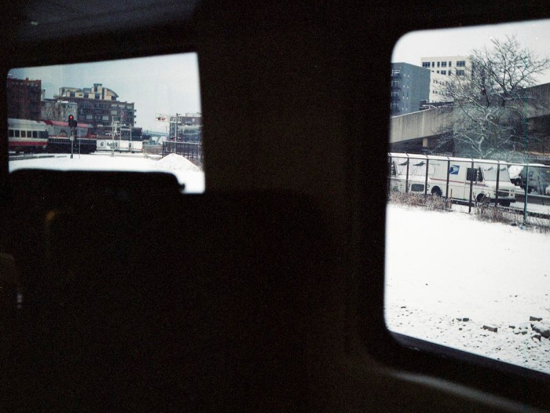 Snow-covered tracks and buildings are visible through two train windows, with vehicles parked along a fence outside.