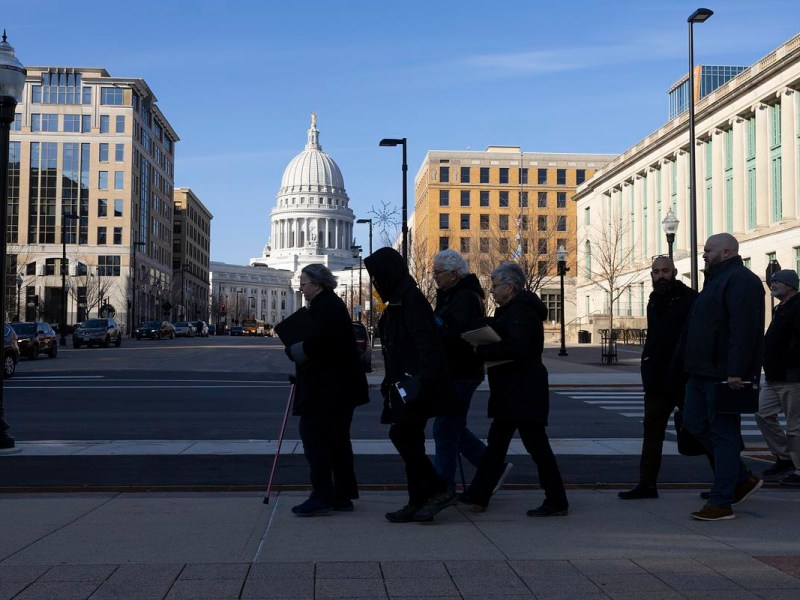 People walk in a line on a sidewalk next to a street, carrying papers, with the Wisconsin State Capitol dome centered in the background between downtown buildings.