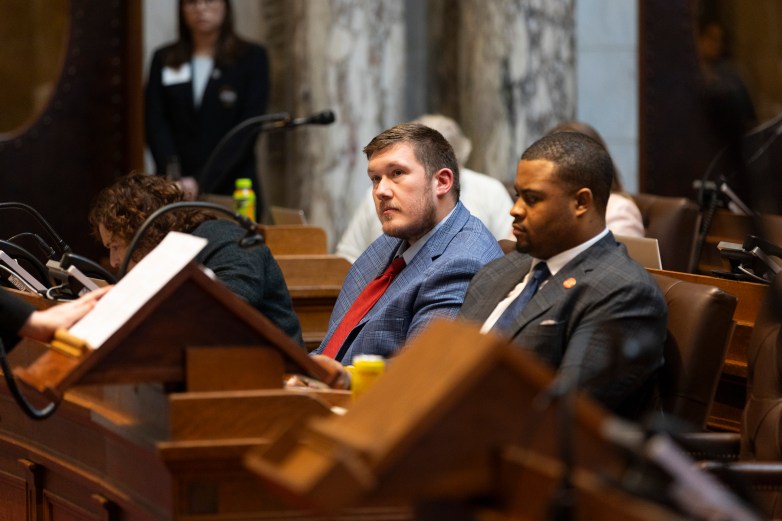 People in suits sit at desks with microphones in a room while a person holds paper at a podium in the foreground.