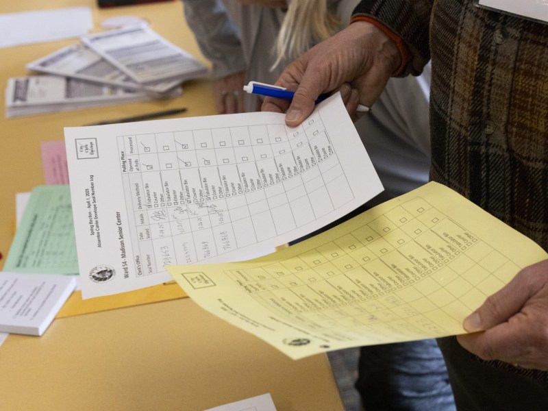 A person holds two pieces of paper, one white and one yellow, and a pen over a table covered with envelopes and other pieces of paper.