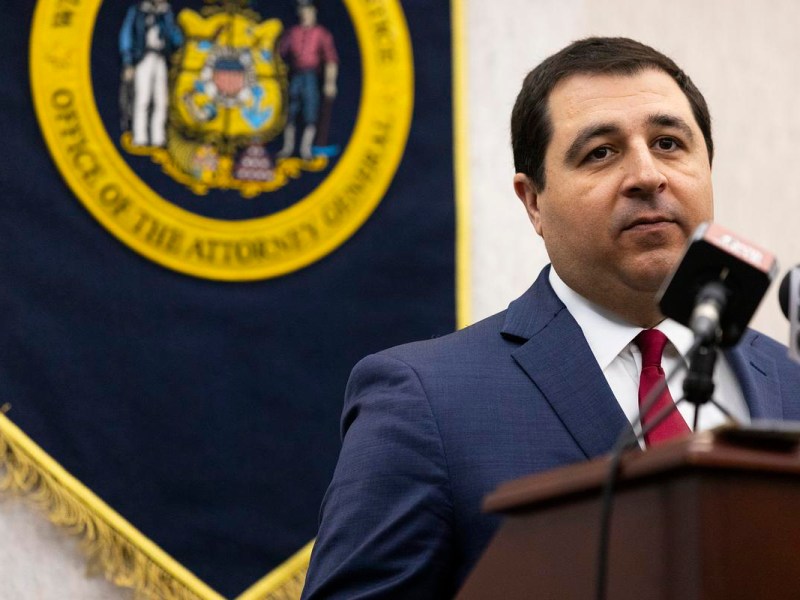 Person in a suit and red tie stands at a podium with microphones. Behind the person is a dark blue banner with the seal of the "Office of the Attorney General"