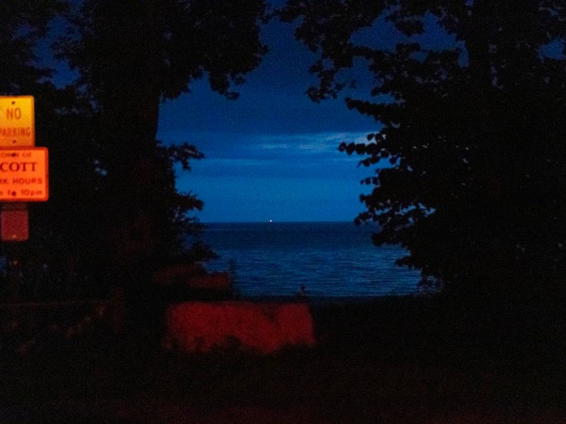 A dark shoreline framed by trees with a faint glow on a “NO PARKING” sign and calm water visible under a deep blue sky.