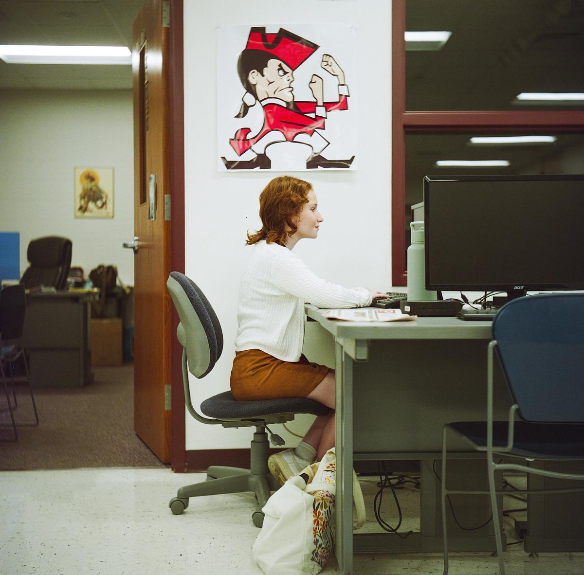 A person sits at a desk with a computer monitor and other items on the desk, with a cartoon poster on the wall behind them.