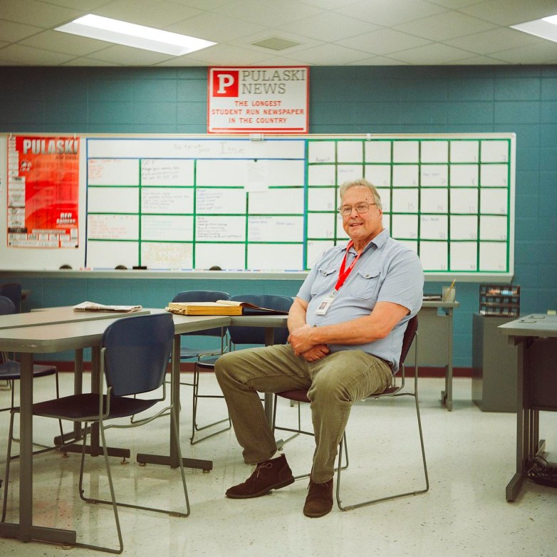 A person wearing a lanyard sits on a chair in a room with desks, a whiteboard and a sign reading “Pulaski News The Longest Student Run Newspaper in the Country.”