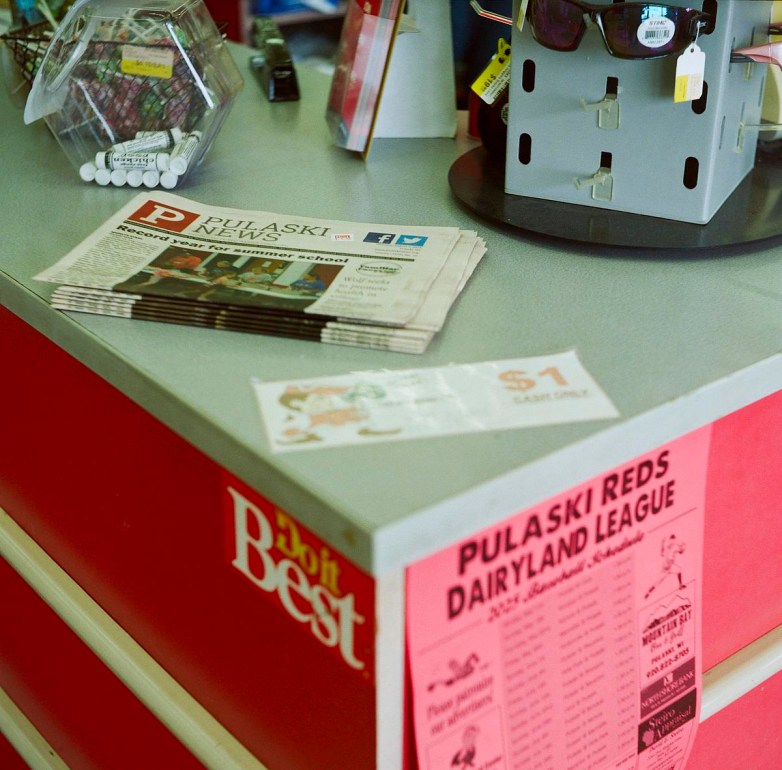 A stack of newspapers, the top one labeled "Pulaski News," on a counter beside a display of sunglasses and a pink flyer for the Pulaski Reds Dairyland League.