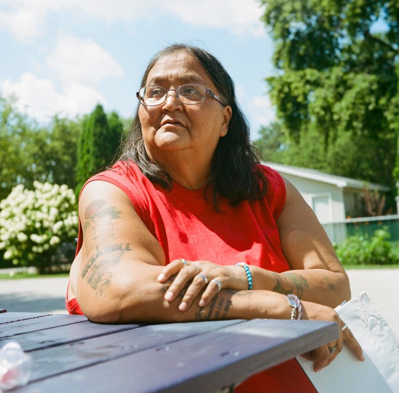 A person wearing glasses and a red sleeveless shirt sits at an outdoor picnic table with trees and a building in the background.