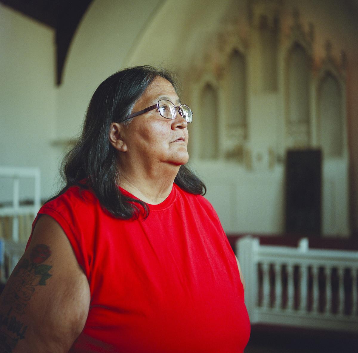 A person wearing glasses and a red sleeveless shirt stands near white railings with out-of-focus arched architectural details in the background.