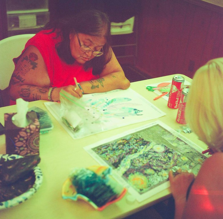 Two people sit at a table working on colorful art projects with craft supplies, a tissue box and drink cans nearby.
