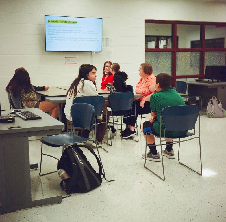 People sit in chairs next to tables with a screen that says “Be specific — provide information” on the wall.