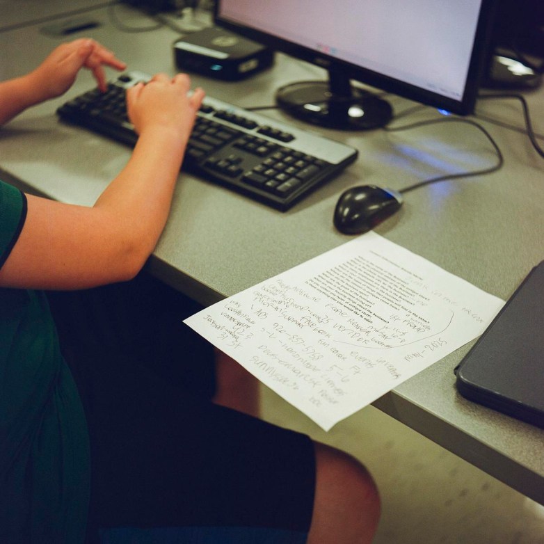 A person types on a computer keyboard at a desk with a piece of paper next to a computer mouse.