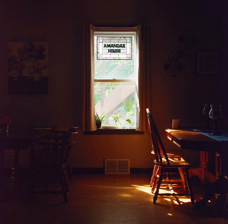 Sunlight shines onto wooden chairs and a table through a window with a stained glass panel reading "AMANDA’S HOUSE."