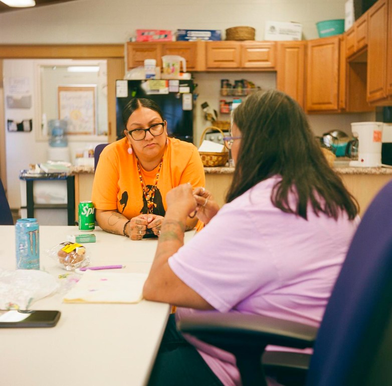 Two people, one wearing an orange shirt and the other a light purple shirt, sit at a table with drinks and craft materials in a kitchen area.
