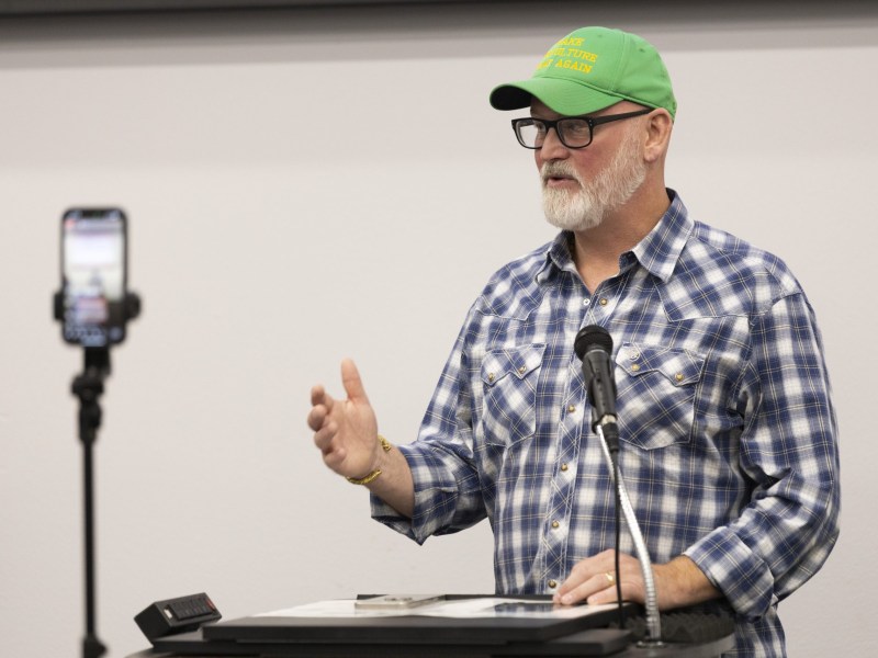 A person wearing a green cap and plaid shirt stands at a podium with a microphone, gesturing with one hand. A phone is on a tripod nearby.