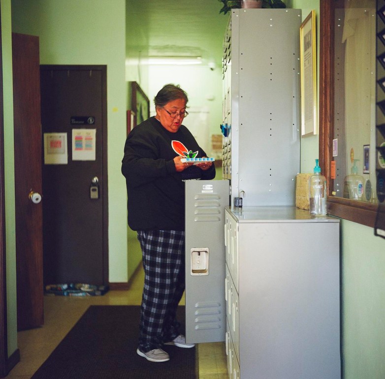 A person stands in a hallway wearing plaid pants and a dark sweatshirt while holding a pill organizer in front of an open locker.