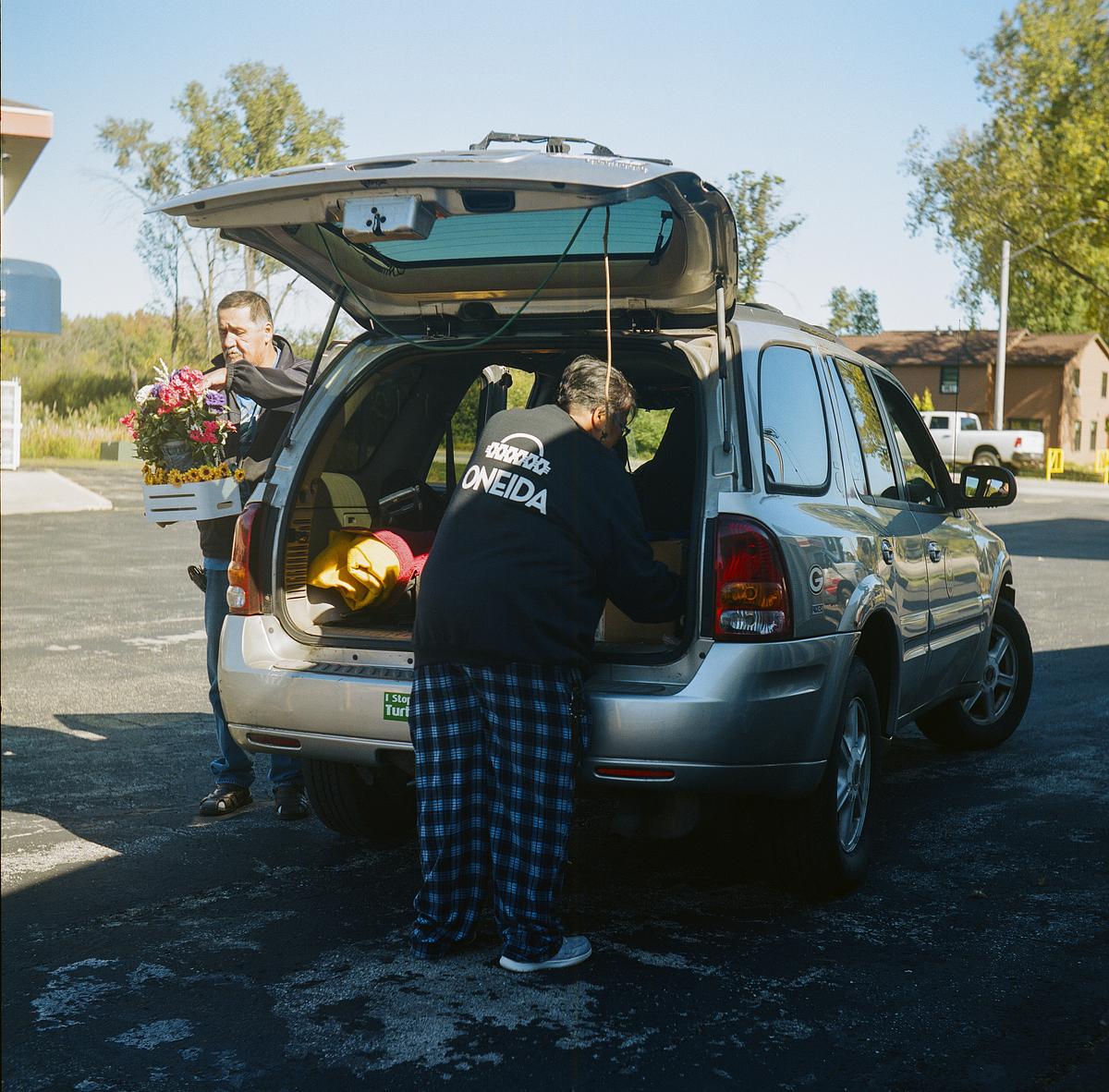 Two people load items into the back of an SUV, one holding a crate of flowers and the other wearing a top with "Oneida" printed on the back.