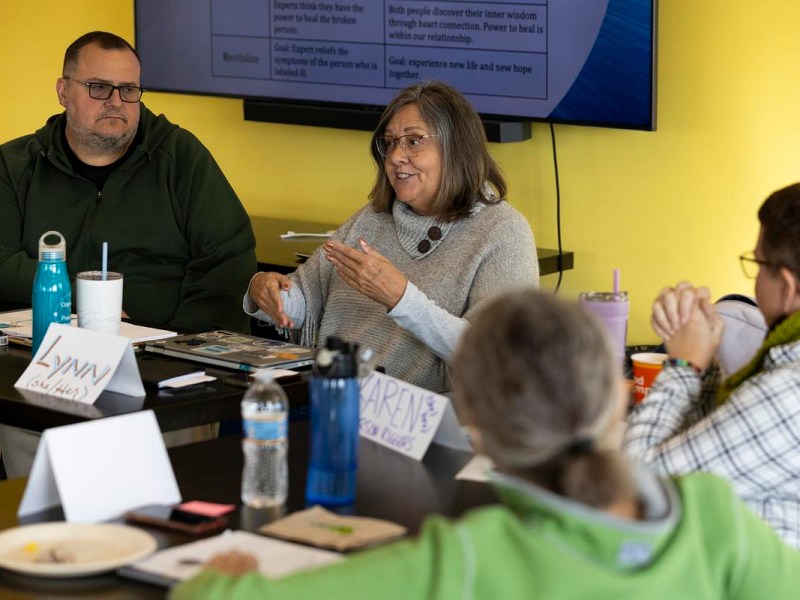 A person gestures while speaking at a table with others, with name cards, notebooks and water bottles visible and a presentation screen showing text in the background.
