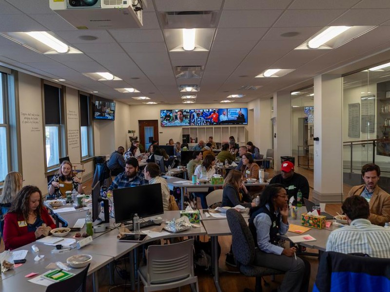People sit at clustered tables in a large room, eating and talking among computers, monitors and papers, with screens on the far wall displaying broadcast images.