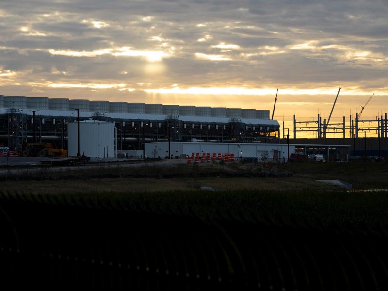 A large industrial building with rows of rooftop units stands behind construction barriers and cranes as sunlight breaks through clouds near the horizon.
