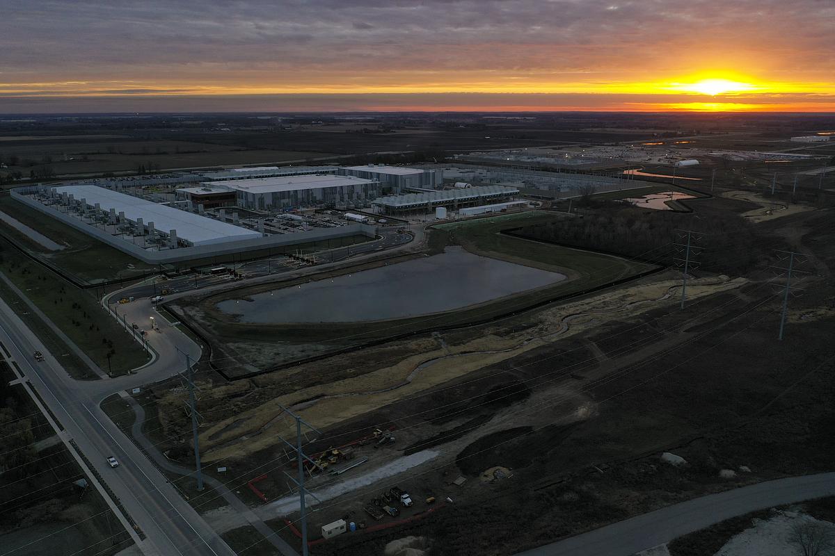 An aerial view of a large industrial complex next to a pond and surrounding construction areas at sunset, with orange light along the horizon under a cloudy sky.