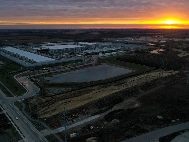 An aerial view of a large industrial complex next to a pond and surrounding construction areas at sunset, with orange light along the horizon under a cloudy sky.