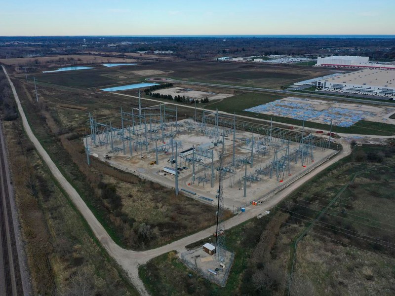 An aerial view of a large electrical facility surrounded by dirt roads, open fields, railroad tracks and nearby industrial buildings