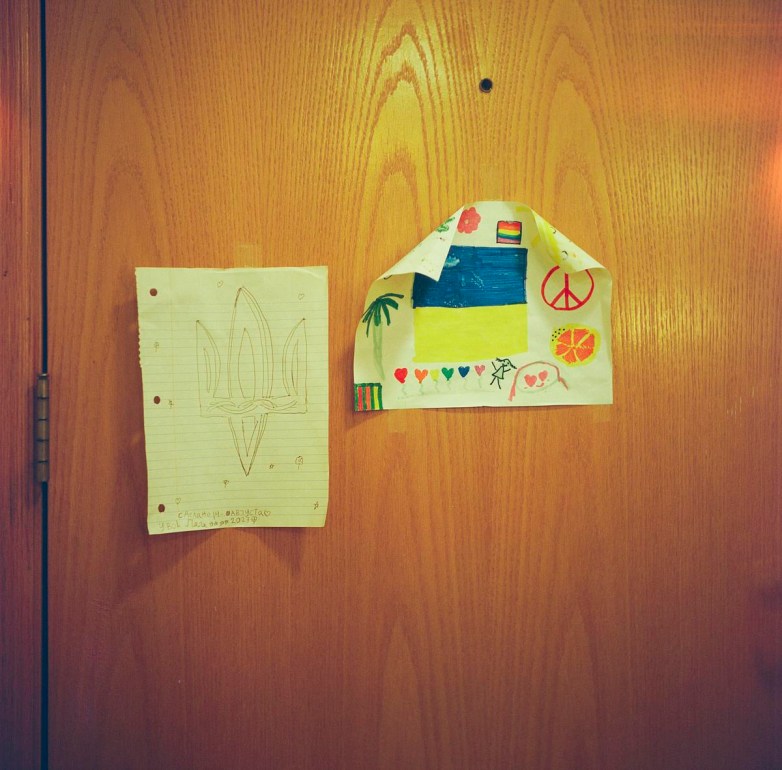 Two drawings are taped to a wooden door, one showing a trident symbol on lined paper and the other a colorful drawing with a blue-and-yellow flag, hearts and peace symbols.