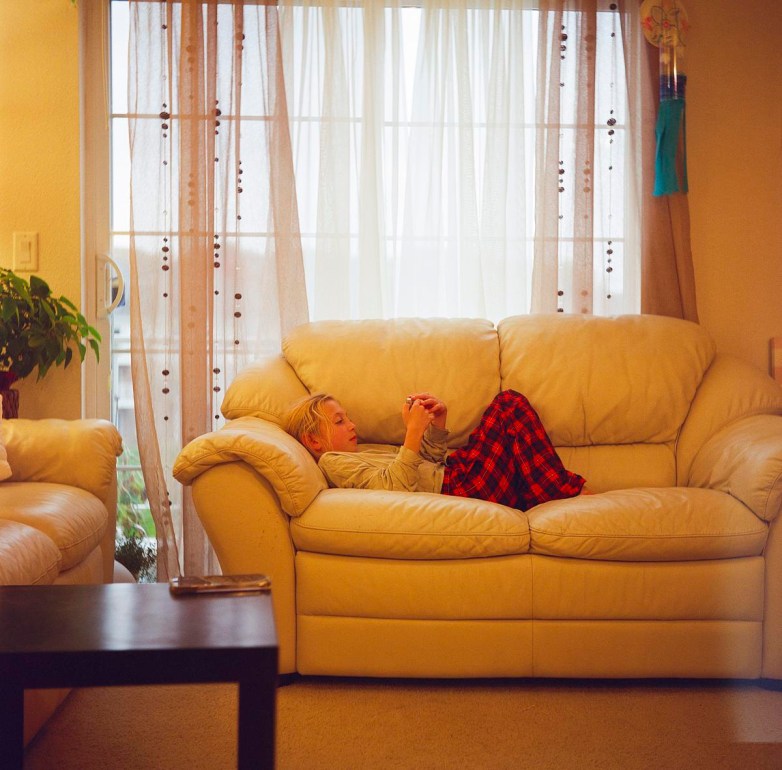 A person lies on a light-colored couch holding a phone, wearing red plaid pants, with sheer curtains and a window behind the couch.