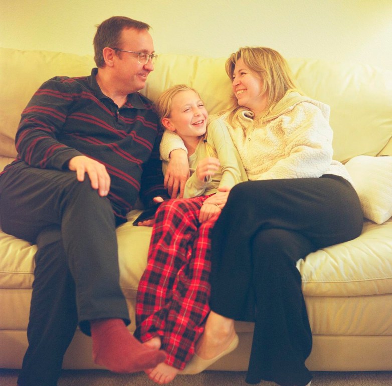 Three people sit close together on a light-colored couch, with one in the middle wearing red plaid pants, while the others look toward each other.