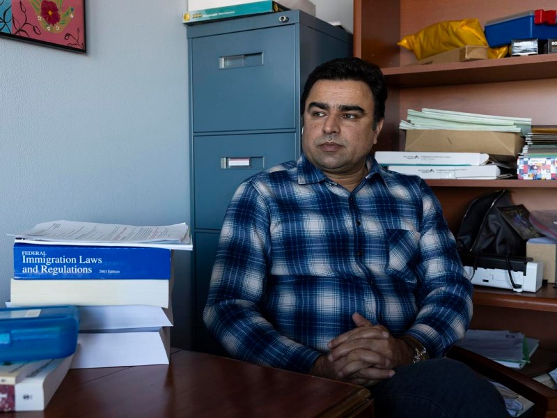 A person sits at a desk in an office, wearing a plaid shirt, with stacks of papers and books including one titled “Federal Immigration Laws and Regulations” nearby.