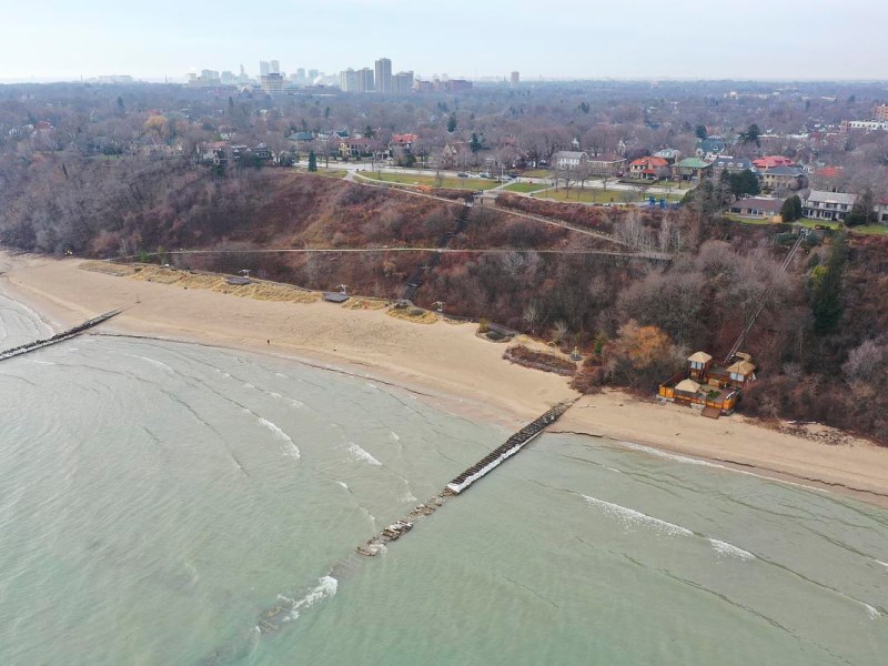 An aerial view shows a sandy beach and greenish lake water with a wooden breakwater, a wooded bluff behind the shore, houses along the top, and a small wooden structure near the sand.