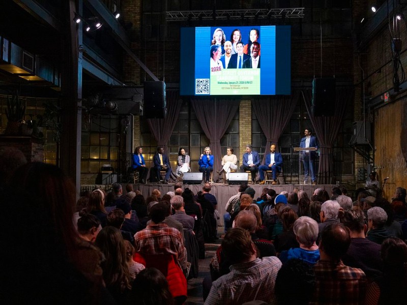 People sit on a stage while a person stands at a podium; a large screen above shows headshots and text reading "2026 Main Street Governor Candidate Forum"
